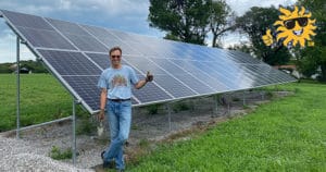 A man in a blue shirt gives the thumbs up in front of a ground-mounted solar panel system newly installed on his property.