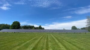 A large array of ground-mounted solar panels on a field with trees in the background.