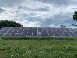 A man stands in front of a large ground-mounted solar panel in a field on a cloudy day.