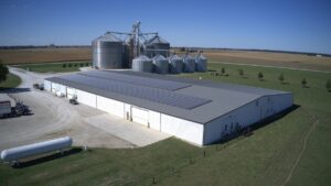 A large warehouse with a plan in the background has a solar panel system installed on its roof.