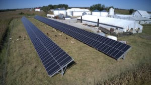 Two large rows of solar panels mounted on the ground outside of an industrial complex.